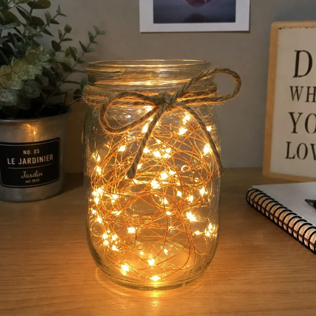 A DIY mason jar centerpiece filled with glowing copper wire fairy lights on a wooden desk